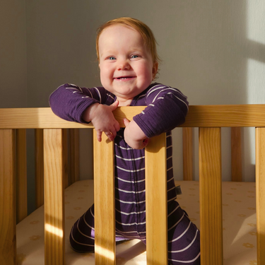 Baby in a purple outfit standing in a wooden crib