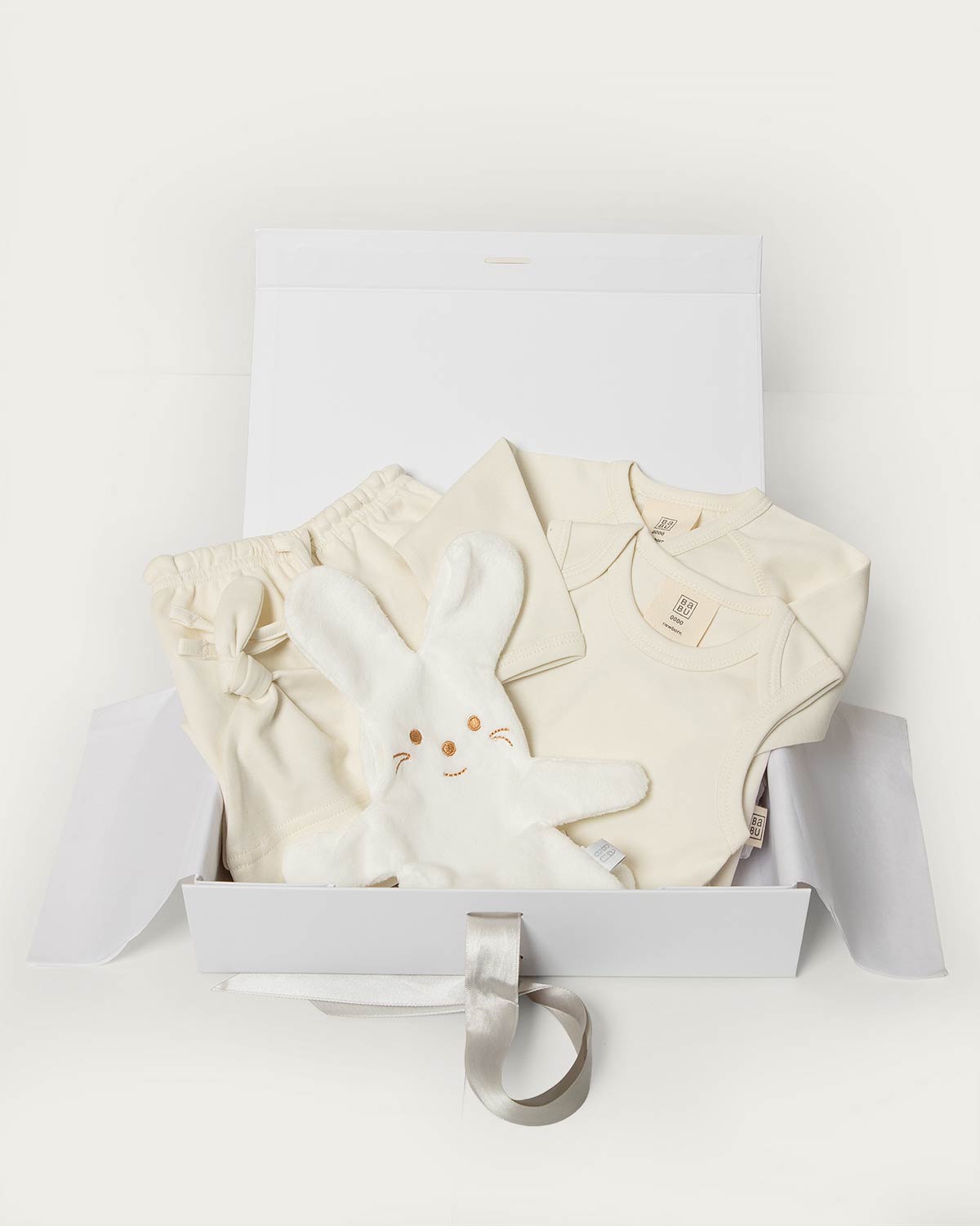 A baby gift set in a white box, featuring a cream-colored romper, matching cream pants, a plush white bunny toy, and a silver ribbon, photographed on a clean white background.