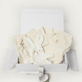 A baby gift set in a white box, featuring a cream-colored romper, matching cream pants, a plush white bunny toy, and a silver ribbon, photographed on a clean white background.