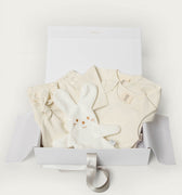 A baby gift set in a white box, featuring a cream-colored romper, matching cream pants, a plush white bunny toy, and a silver ribbon, photographed on a clean white background.
