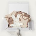 A baby gift set in a white box, featuring a beige and brown clothing set, a white plush bunny toy, and a white gift box with a ribbon.
