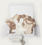 A baby gift set in a white box, featuring a beige and brown clothing set, a white plush bunny toy, and a white gift box with a ribbon.