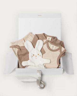A baby gift set in a white box, featuring a beige and brown clothing set, a white plush bunny toy, and a white gift box with a ribbon.