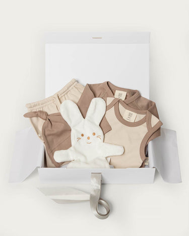 A baby gift set in a white box, featuring a beige and brown clothing set, a white plush bunny toy, and a white gift box with a ribbon.