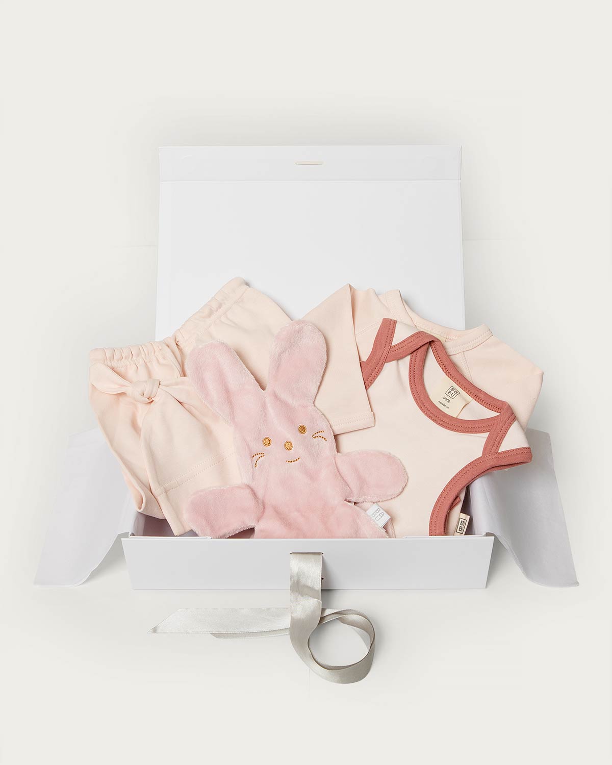 A baby gift set in a white box, featuring a light pink plush bunny toy, a pale pink bodysuit with dusty rose trim, matching shorts, and a white satin ribbon, on a clean white background.