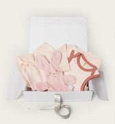A baby gift set in a white box, featuring a light pink plush bunny toy, a pale pink bodysuit with dusty rose trim, matching shorts, and a white satin ribbon, on a clean white background.