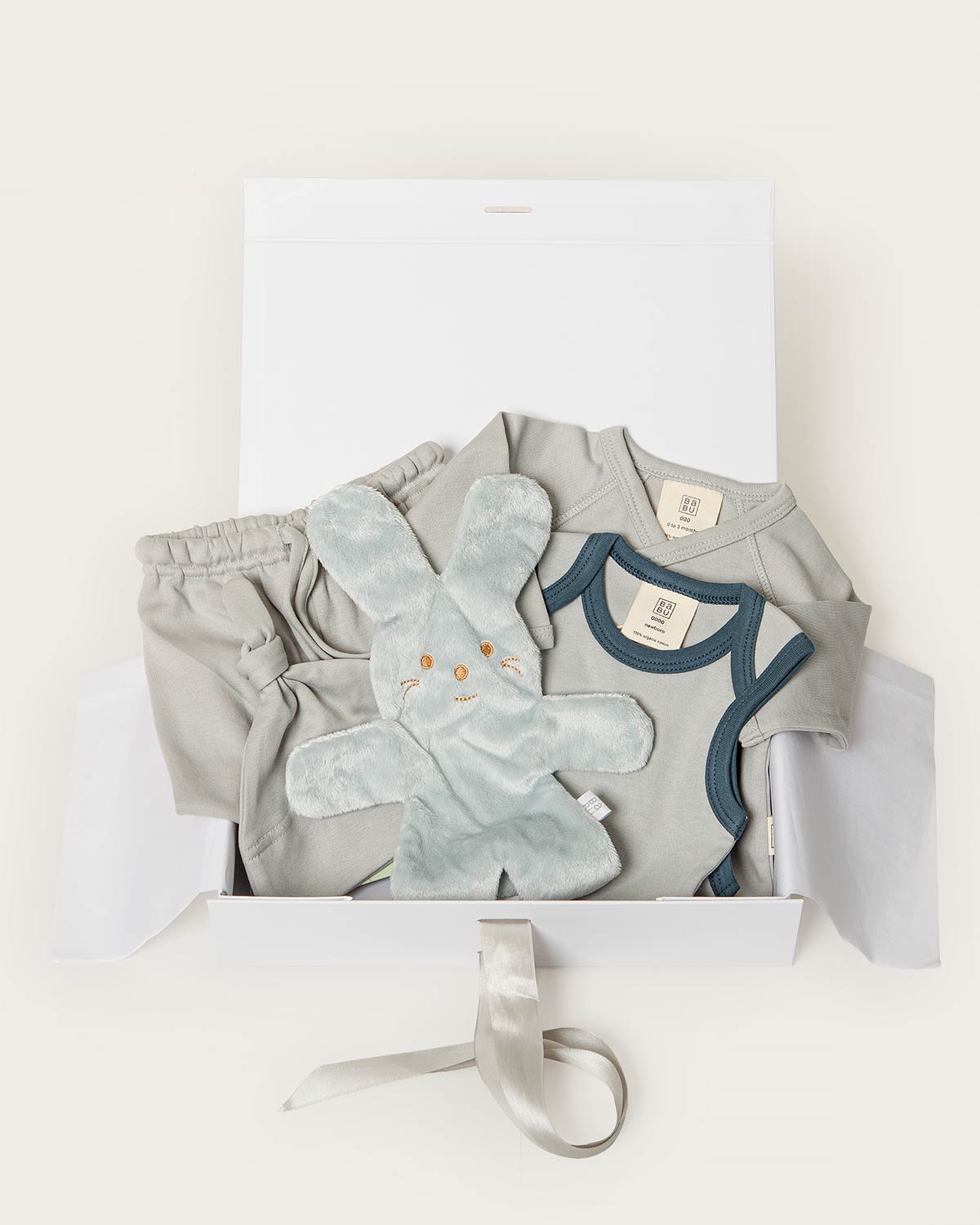 A baby gift set in a white box, featuring a light blue short-sleeve bodysuit with navy trim, a matching pair of light blue pants, a plush light blue bunny toy, and a white box with a gray ribbon.