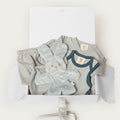 A baby gift set in a white box, featuring a light blue short-sleeve bodysuit with navy trim, a matching pair of light blue pants, a plush light blue bunny toy, and a white box with a gray ribbon.