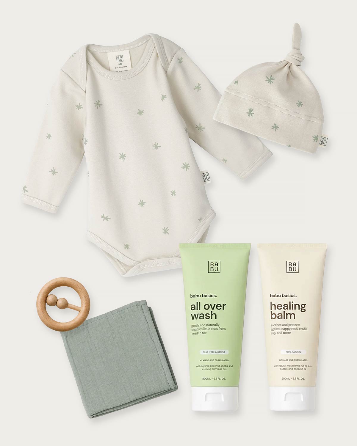 Baby clothing and care set on a light neutral background, including a long-sleeve bodysuit, knotted beanie, tubes of Bath & Body all over wash and healing balm, a wooden teething ring, and a folded cloth.
