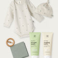 Baby clothing and care set on a light neutral background, including a long-sleeve bodysuit, knotted beanie, tubes of Bath & Body all over wash and healing balm, a wooden teething ring, and a folded cloth.