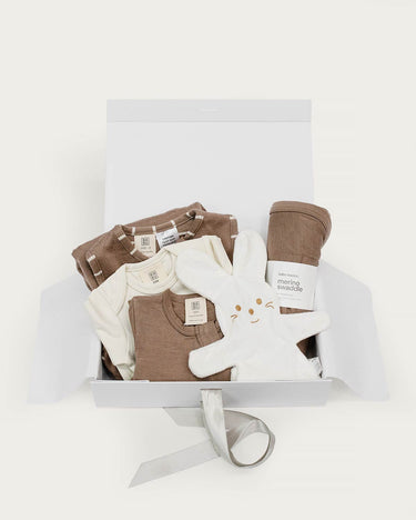 A baby gift set arranged inside an open white box on a clean white background. The set includes brown merino sleeping bag,  brown footless onsie and a cream baby gown, a brown merino swaddle, and a white bunny toy.