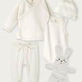 A 4-piece baby clothing set in soft off-white/cream, including a long-sleeve wrap top, sleeveless bodysuit, footed pants with a drawstring, and a knotted beanie, arranged with a plush bunny toy on a light gray background.