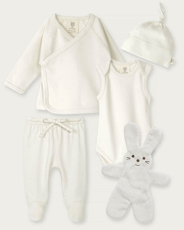A 4-piece baby clothing set in soft off-white/cream, including a long-sleeve wrap top, sleeveless bodysuit, footed pants with a drawstring, and a knotted beanie, arranged with a plush bunny toy on a light gray background.