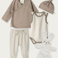 A baby clothing set laid out on a light neutral background, including a taupe wrap shirt, cream bodysuit with taupe trim, cream footed pants, a taupe knotted beanie, and a white plush bunny toy.