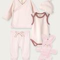 A 5-piece baby clothing set in pale pink, including a long-sleeve wrap top, sleeveless bodysuit, footed pants, a knotted beanie, and a plush bunny toy, arranged on a light gray background.