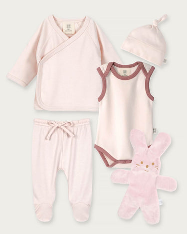 A 5-piece baby clothing set in pale pink, including a long-sleeve wrap top, sleeveless bodysuit, footed pants, a knotted beanie, and a plush bunny toy, arranged on a light gray background.