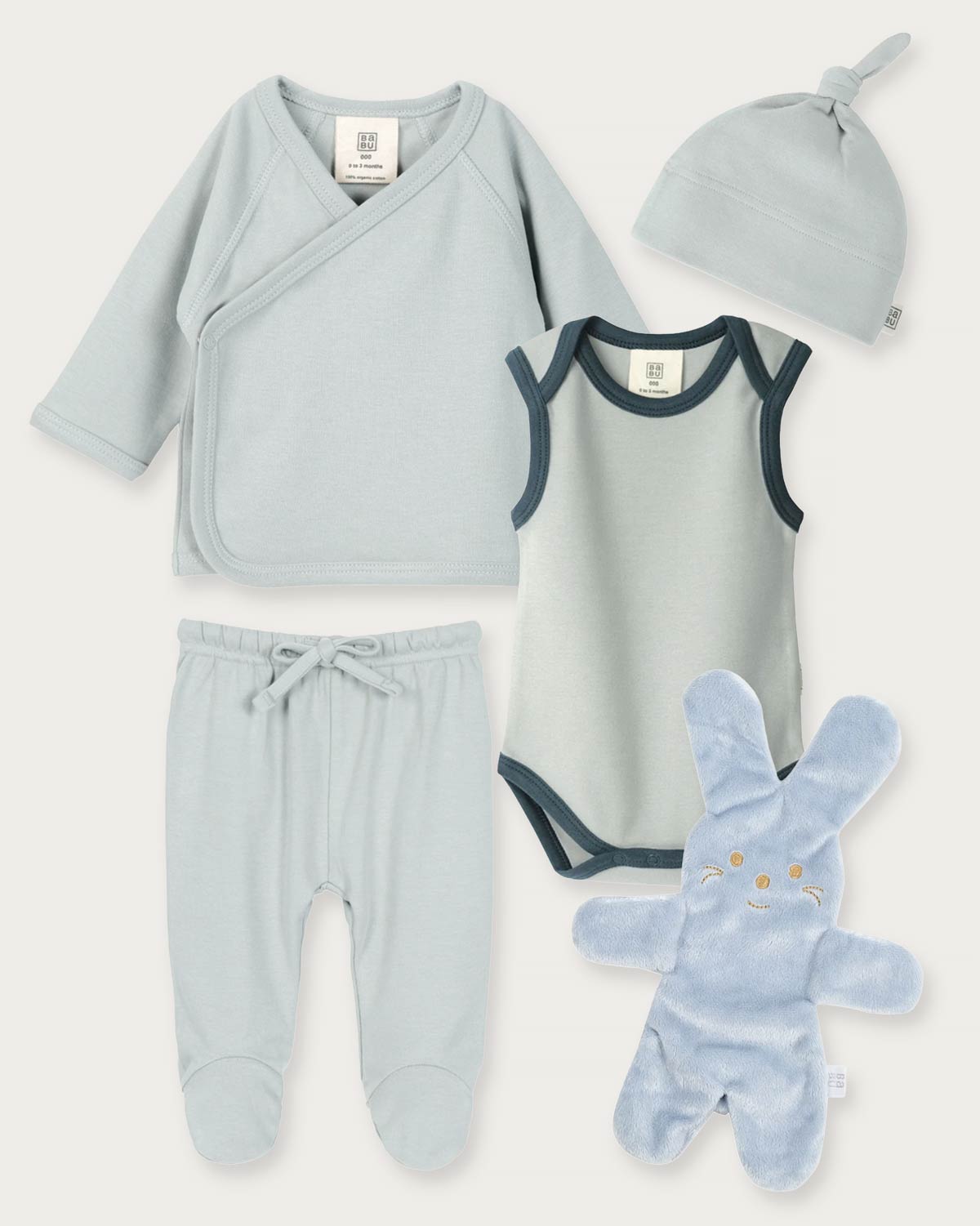 A baby clothing set laid out on a light beige background, including a pale blue long-sleeve wrap shirt, matching footed pants, a pale blue bodysuit with dark trim, a knotted beanie, and a plush bunny toy.