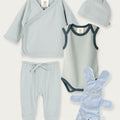 A baby clothing set laid out on a light beige background, including a pale blue long-sleeve wrap shirt, matching footed pants, a pale blue bodysuit with dark trim, a knotted beanie, and a plush bunny toy.